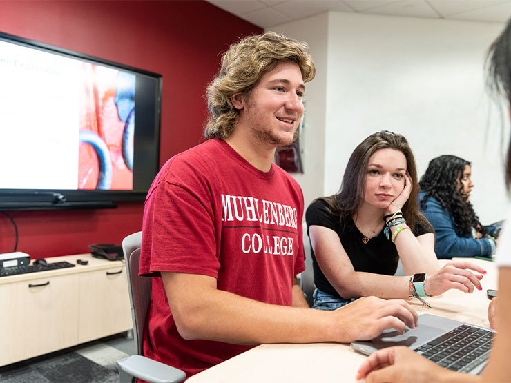 A pair of young adults seated in front of an open laptop speak to a person who is out of frame of the camera.