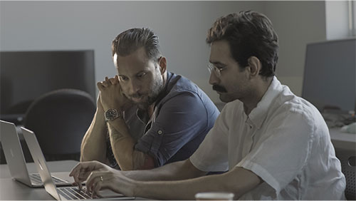 A video still image of a pair of college instructors sit alongside each other at an open laptop computer.