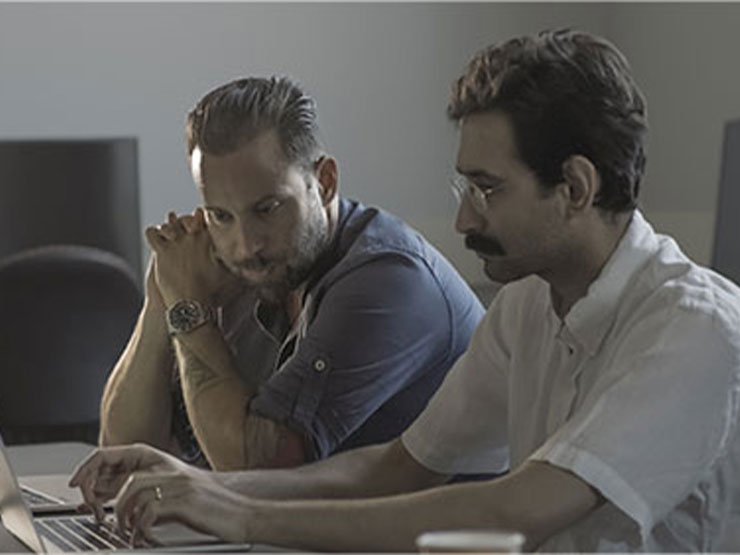 A video still image of a pair of college instructors sit alongside each other at an open laptop computer.