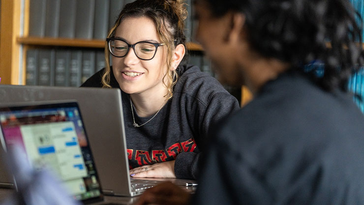 Two young adults chat while working on laptop computers in a room with bookshelves lining the walls.