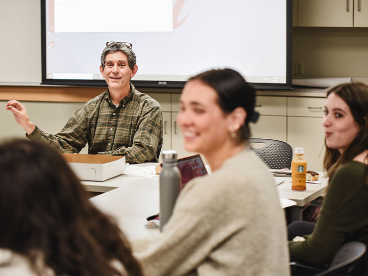 College students in a classroom grin and look off-camera as a professor speaks.