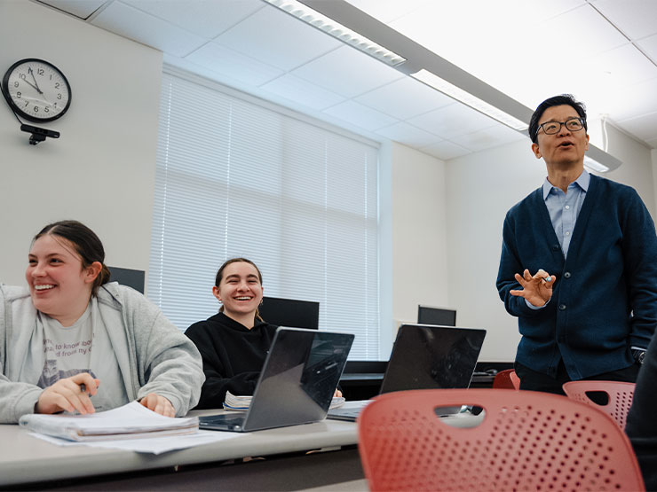 A pair of students smile and chuckle while listening to a college professor speak in a classroom.