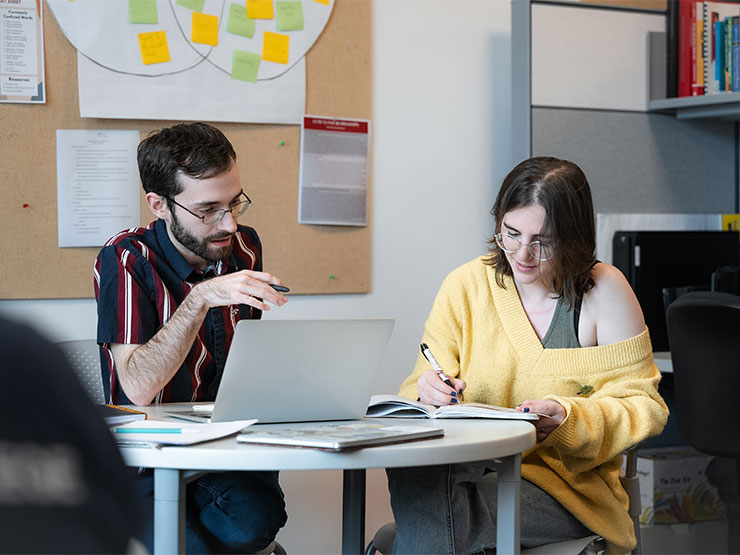 A pair of college students sit together a table in the library writing center with an open laptop and notebook.