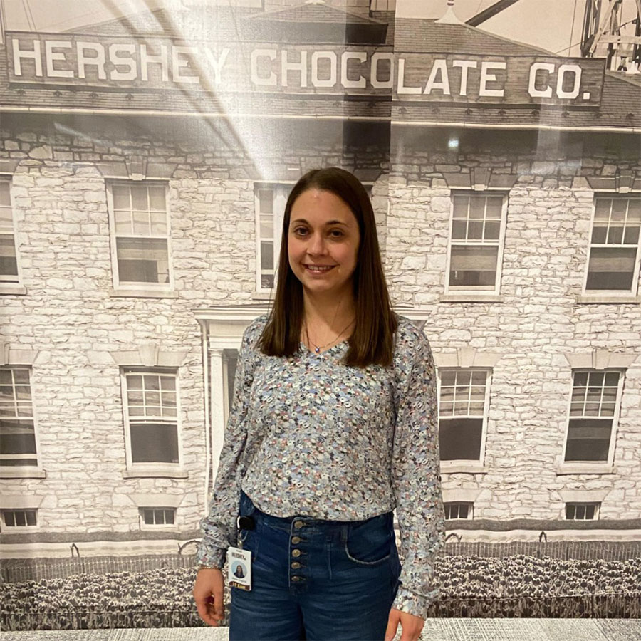 A young adult in a grey, patterned blouse stands in front of a wall adorned with an old photo of a building that says 