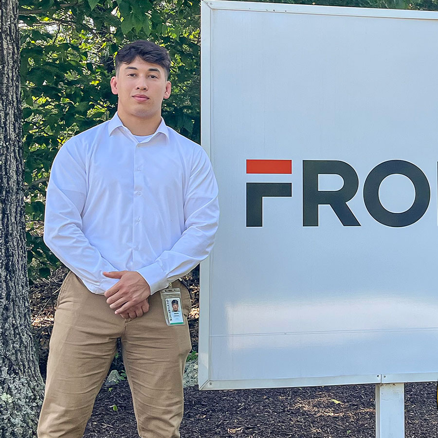 A young adult in a white dress shirt and brown slacks stands next to the sign of a business.