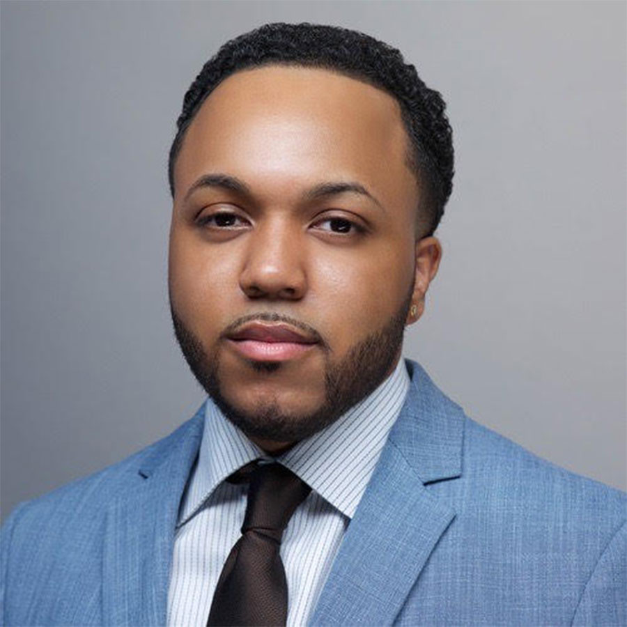 An adult with facial hair, a patterned dress shirt, black tie and blue suit jacket looks to the camera.