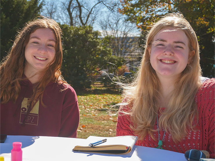 A pair of college students sit in the sunny outdoors at a table during an event