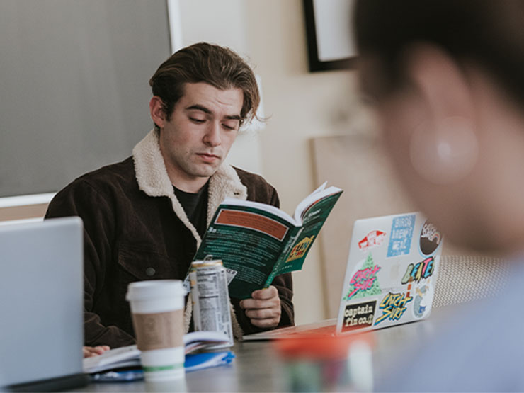 Students sit around a desk, one of them in a brown jacket, reads a paperback book.