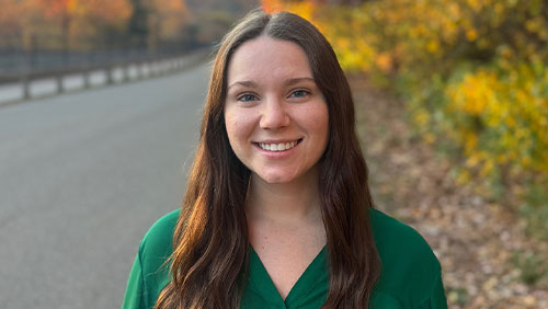 A young person in a green shirt smiles for a photo outside with fall foliage in the background