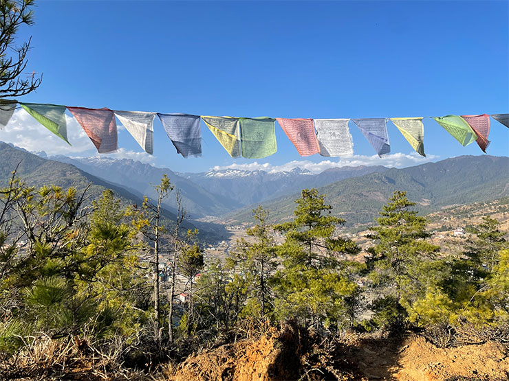 A colorful prayer flag, suspended over a scenic pass with snow-capped mountains in the distance, dances in the wind.