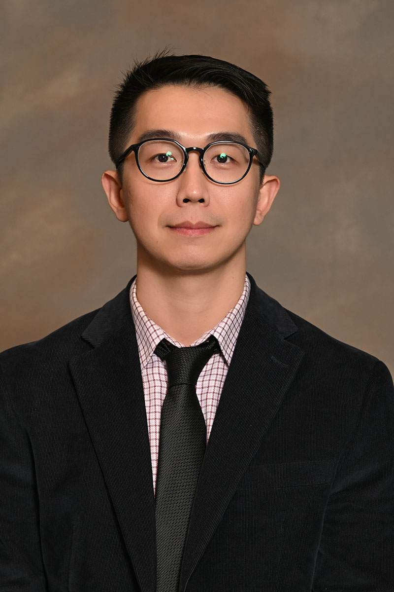 Professor Anh Sy Huy Le standing in front of bookshelf in office setting wearing glasses and collared shirt.