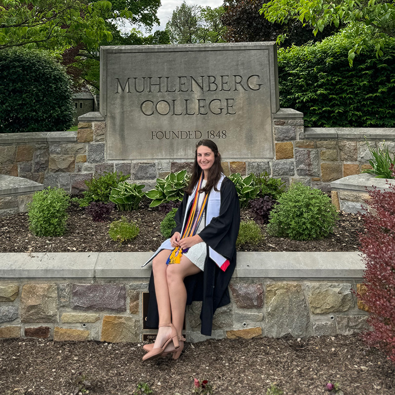 Muhlenberg History program alumni, Sophia Framm '24, seated outdoors on stone campus signage reading 