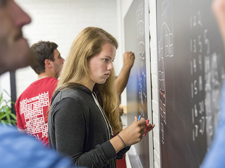 College students in a classroom stand a chalkboard and solve formulas.