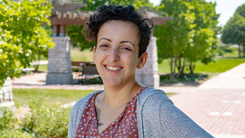 A headshot taken outside of an adult with short dark hair and is wearing a flowered top and gray sweater