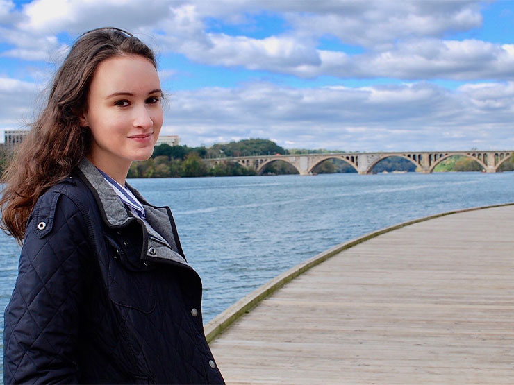 A college student smiles at the camera while standing alongside a river.