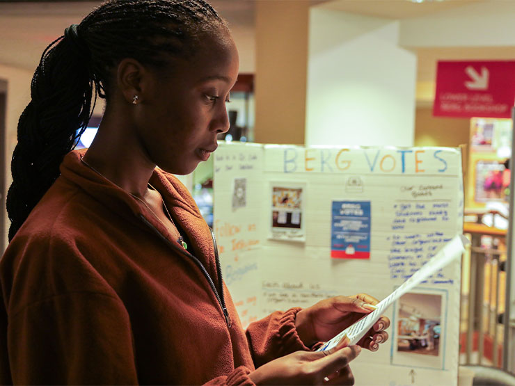 A college student looks thoughtfully at a paper held in their hands, a table behind them contains info on a club called 