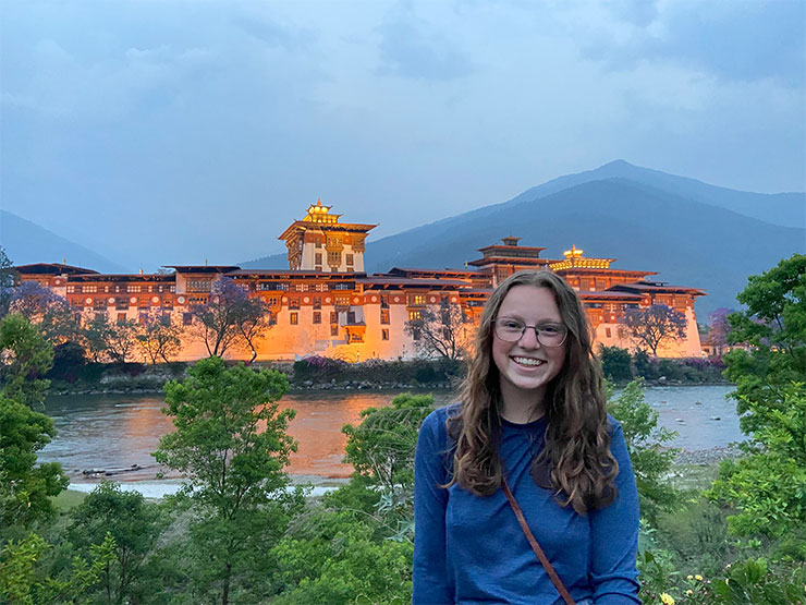 A young woman with long, wavy brown hair and glasses smiles while standing on a riverbank, a large Buddhist monastery in the distance.