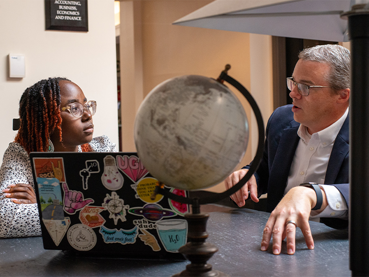 A student and instructor discuss research at a table with an open laptop and globe.