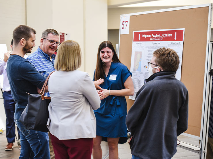 A college student in a blue dress laughs while speaking with a group gathered around a research poster.