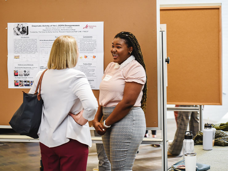 A college student smiles widely while speaking with an adult about a research poster.