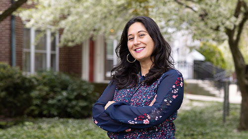 An adult with shoulder-length black hair, large hoop earrings and a dark blue floral dress stands outdoors on a college campus, arms crossed and smiling.