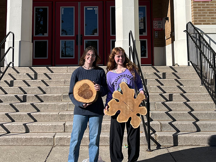 A pair of college students stand side-by-side holding large cross sections of tree trunks in front of an elementary school.