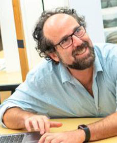 An adult in a light blue shirt smiles while working at a desk.