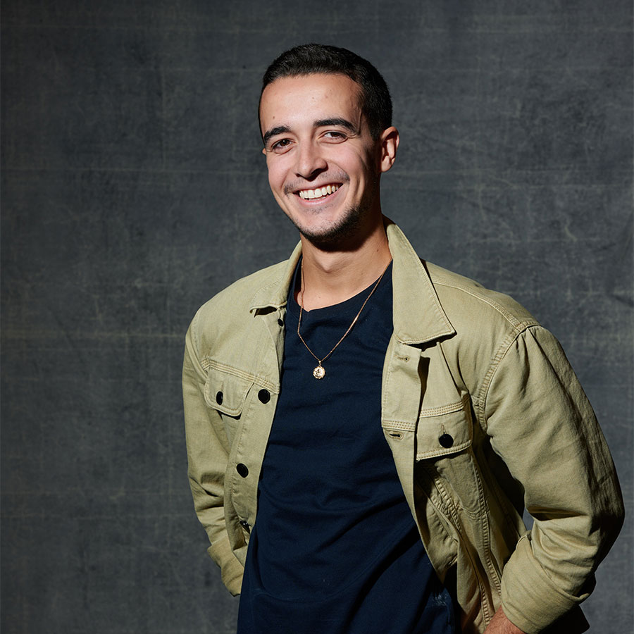 A professional headshot of a young adult smiling in an olive green jacket and black shirt.