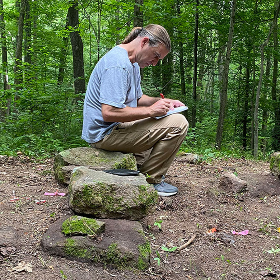 A man, seated on moss-covered stones in a forest, writes in a notebook perched on his knee.