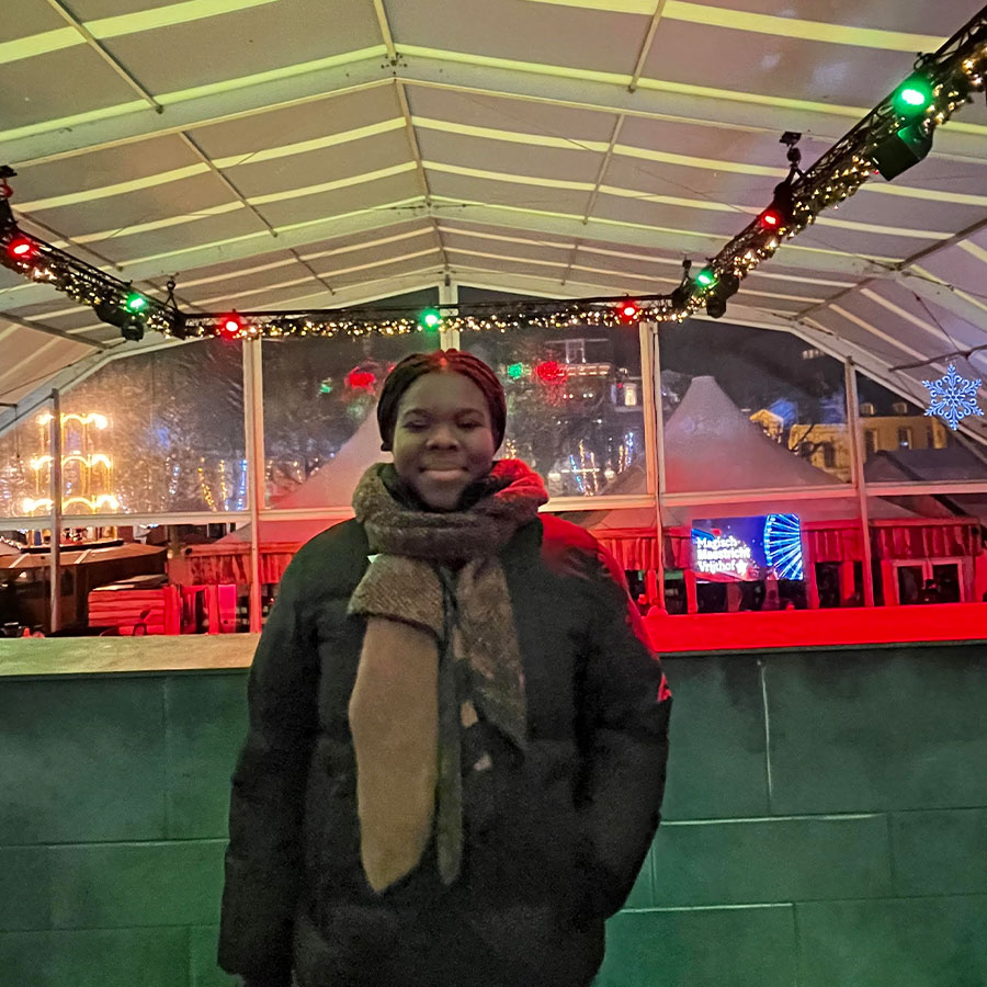 A college students dressed in a winter coat and scarf smiles while standing under an overhang decorated with festive holiday lights.