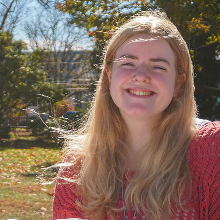 A college student with long blonde hair grins at the camera while seated outdoors in the sunshine