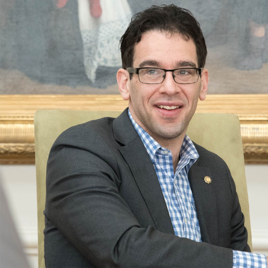 A man in a suit and glasses smiles while sitting at a table in front of a large painting.