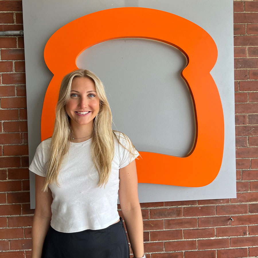 A young adult with long blonde hair smiles while standing next to an orange logo shaped like a piece of toast.