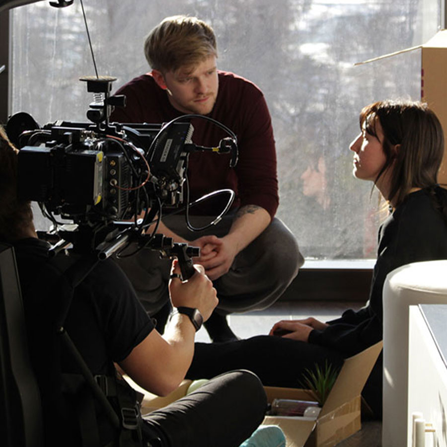 Two actors perform in front of a video camera with a big window overlooking a city behind them.