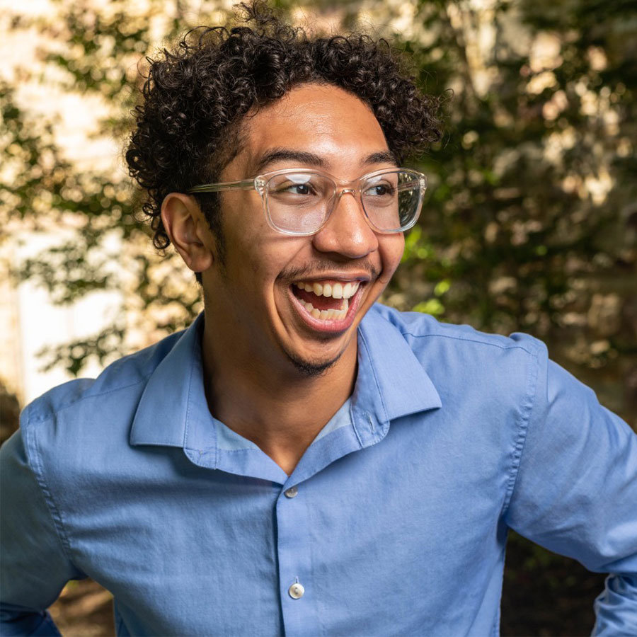 A young Black man with glasses wears a light blue dress shirt and grins widely outdoors.