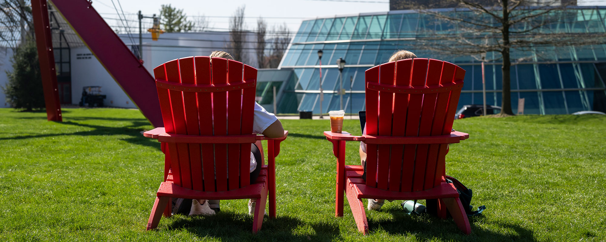 Two people sit in a pair of red Adirondack chairs that are facing away from the camera on a college campus.