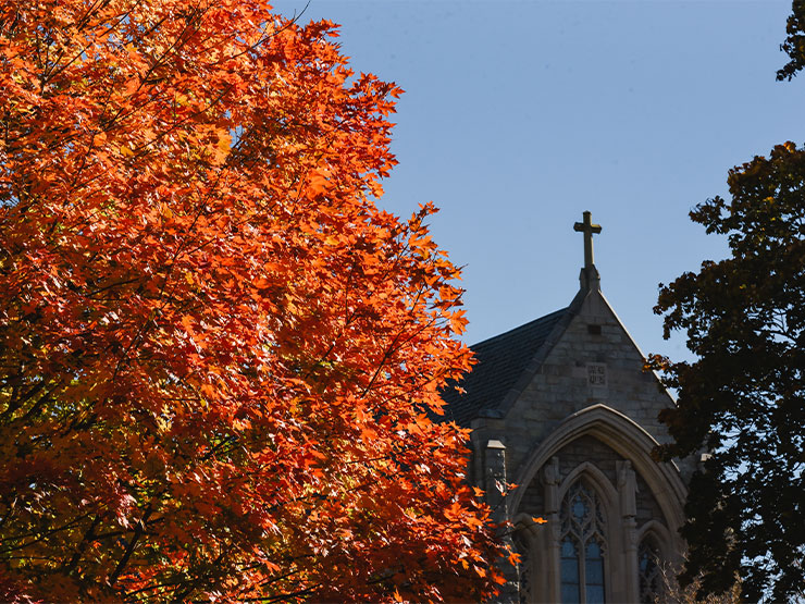 The bright orange foliage of a tree stands against a blue sky with the spire of Egner Memorial Chapel in the distance.