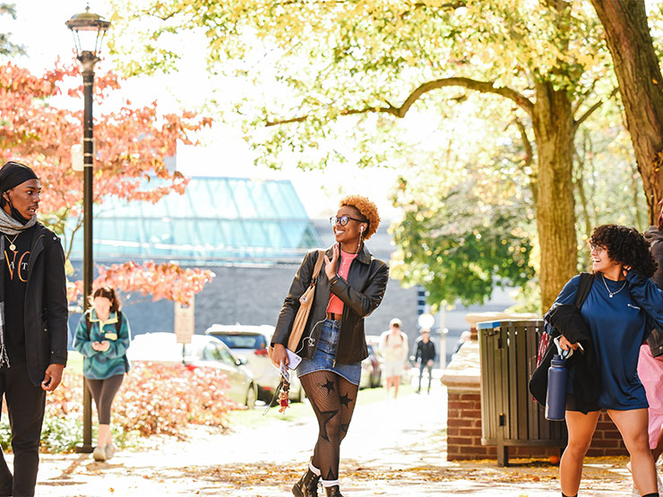 A pair of students wave to a third as they go separate ways on a college campus under a bright canopy of autumn leaves.