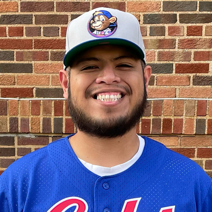 Muhlenberg College alumni smiling positioned in front of red brick wall wearing a white visor hat with blue Cubs jersey.