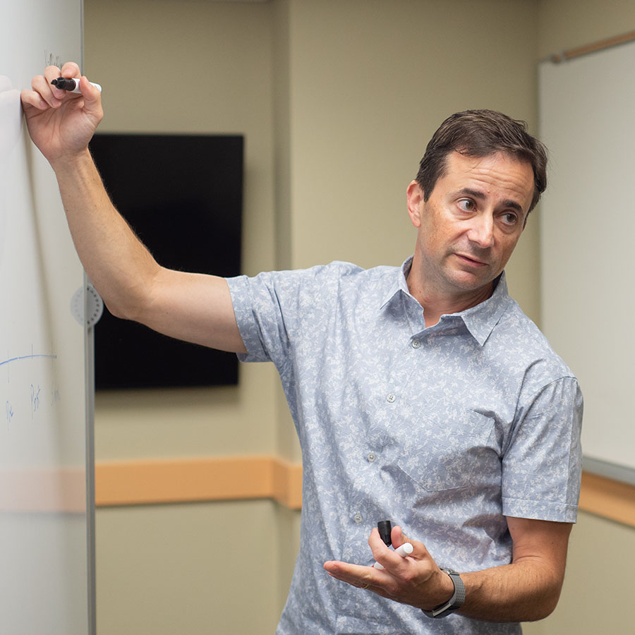 Faculty member Mark Scuitto wearing collared shirt standing at whiteboard holding marker.