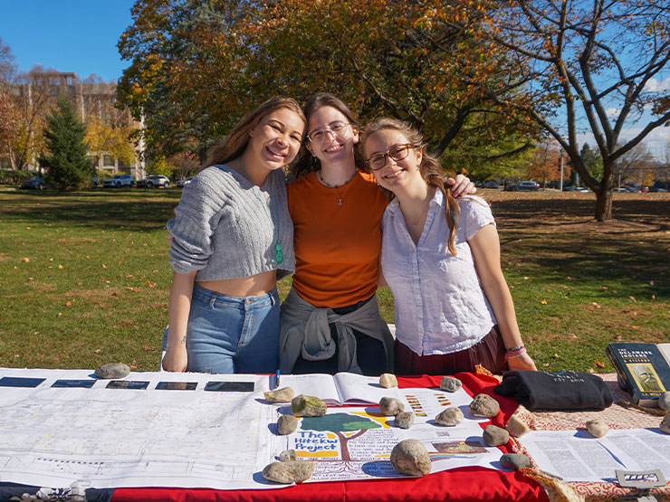 A trio of college students lean close to one another for a group photo outdoors behind a table filled with materials and information.