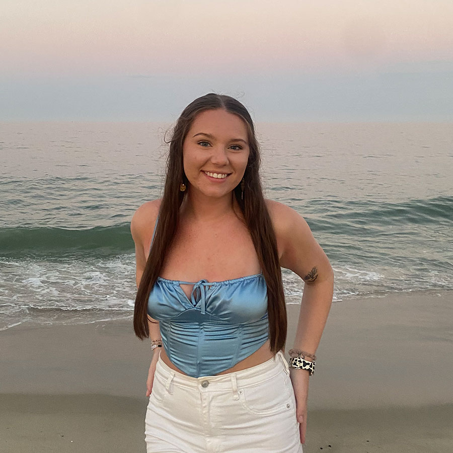 A smiling young woman stands, hands in back pockets, on the shore of a beach.