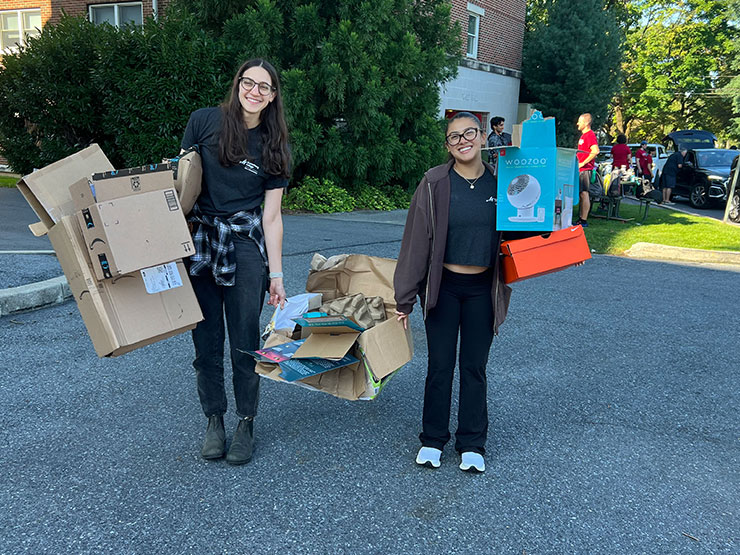 A pair of college students smile for the camera while carrying boxes of cardboard during move-in day.