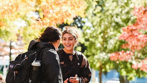 Two young adults chat outdoors, surrounding by bright fall foliage.