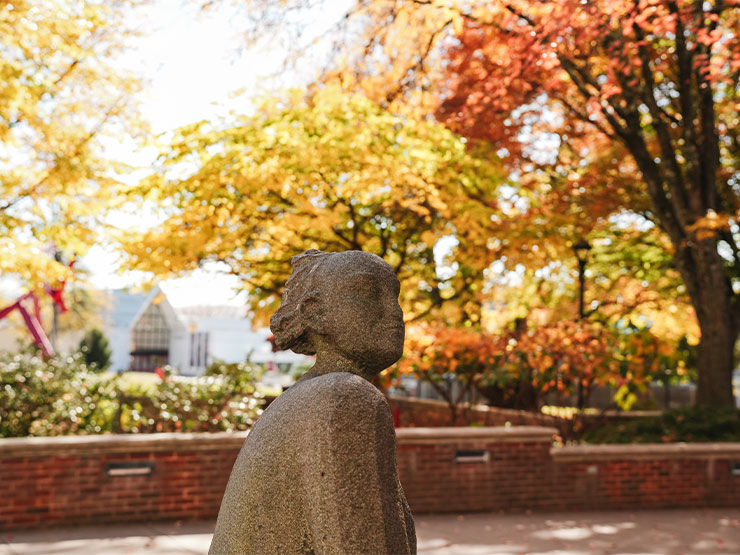 A stone statute of a figure looking into the distance stands in plaza flanked by trees with colorful fall foliage.