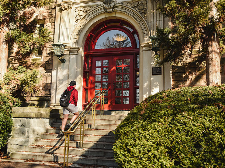 A student wearing a red sweatshirt, shorts and a black backpack walks up the stairs to the red door of Ettinger Hall on the campus of Muhlenberg College.