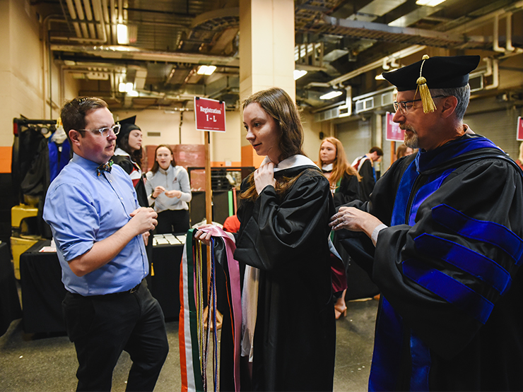 Two adults help a young adult in commencement regalia get dressed ahead of the ceremony.