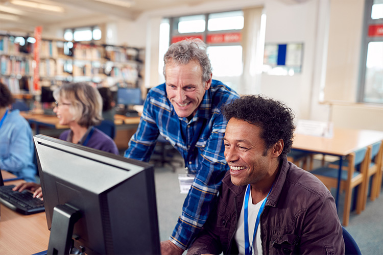 Student and mentor in discussion at computer desk in library setting.