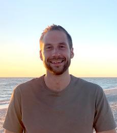 A bearded man smiles at the camera with a large body of water behind him.