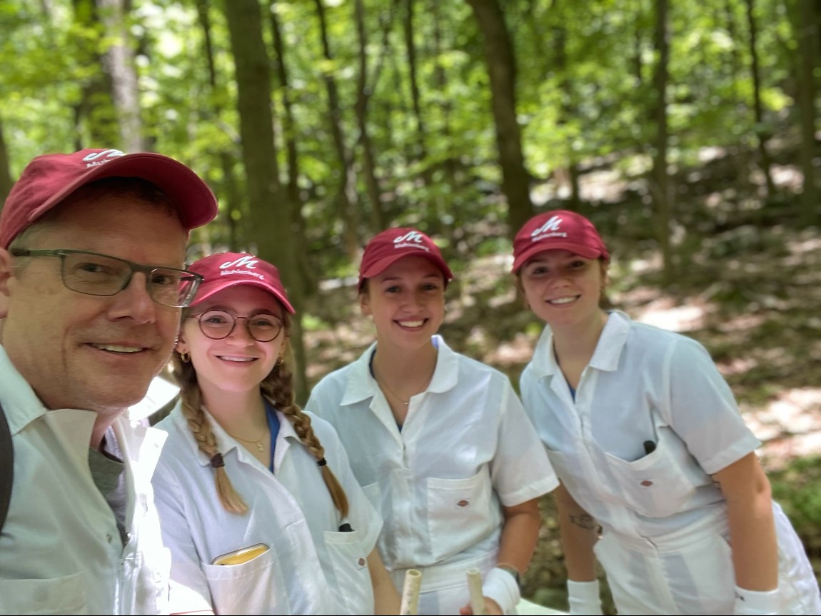 A man and three students stand in a forested area.